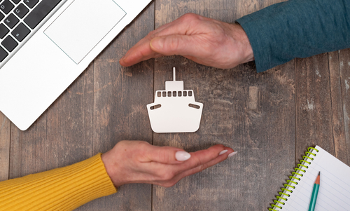 Two hands reach toward a paper ship model on a wooden table beside a laptop and notepad.