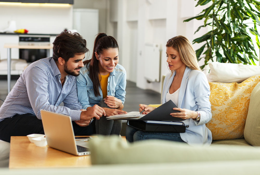 Three young adults discuss documents together in a bright living room with plants and a laptop.