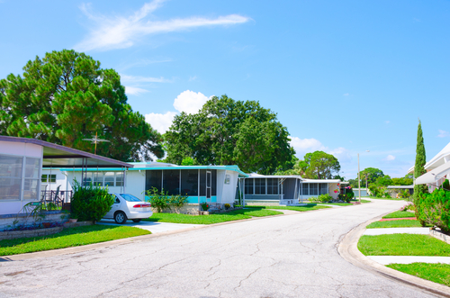 Sunlit street in a residential area with mobile homes and lush green trees.