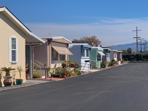 Row of colorful mobile homes with gardens along a paved road under a clear blue sky.